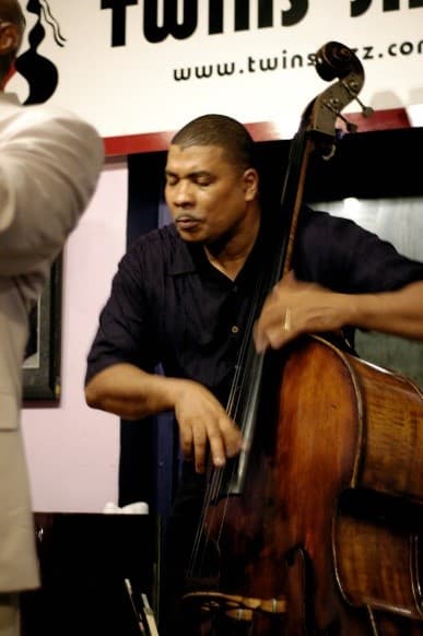 A bassist plays at the Twins Jazz club, with the club's sign on the wall behind him.