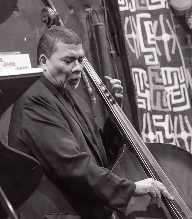 A black-and-white photo of a bassist playing in front of a patterned textile wall hanging.