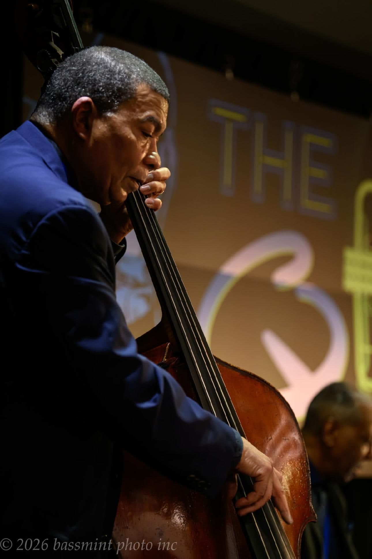 A bassist in a blue jacket plays the upright bass with his eyes closed, lit warm against a glowing sign, with a pianist in the background.