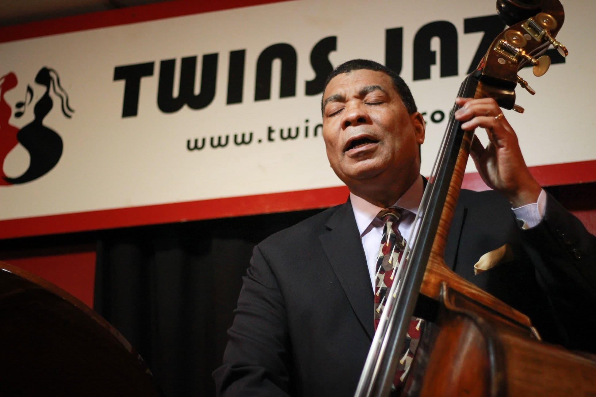 A bassist in a dark suit sings along with his eyes closed as he plays the upright bass, in front of a Twins Jazz banner.