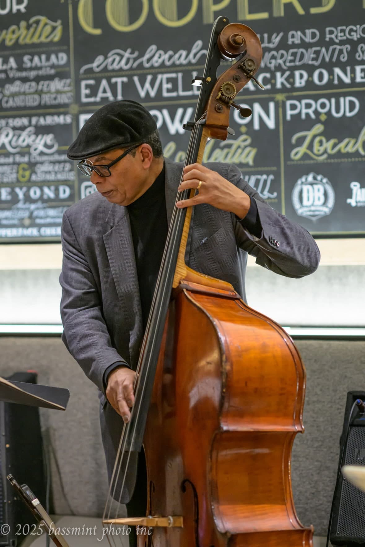A bassist in a flat cap, glasses and a grey jacket plays the upright bass in front of a chalkboard restaurant menu.