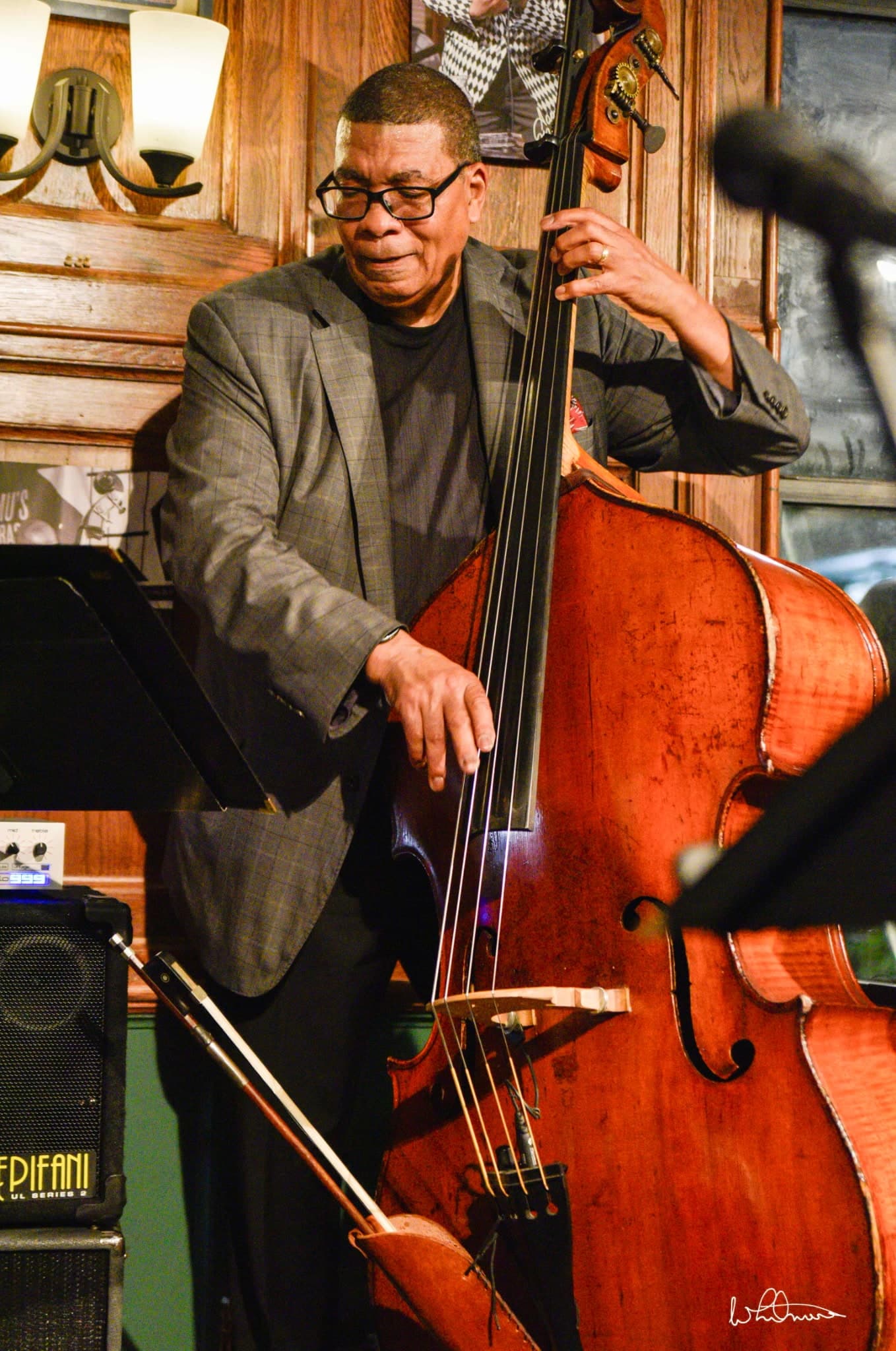 A bassist in glasses and a grey checked jacket smiles as he plays the upright bass in a wood-paneled room, beside a wall lamp.