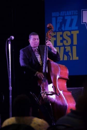 A bassist performs on stage in front of a Mid-Atlantic Jazz Festival banner.