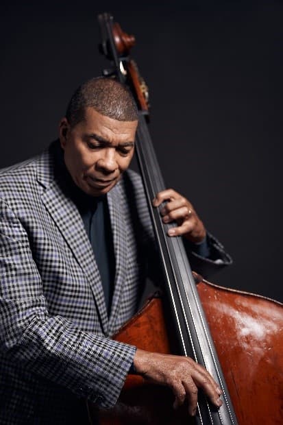 A bassist in a checked jacket plays the upright bass against a dark studio backdrop, looking down at the strings.