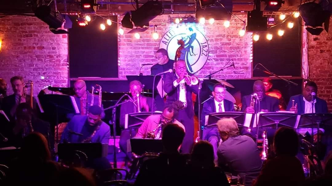A big band performs under the Blues Alley sign and strings of stage lights, a trumpeter soloing at the front while the horn sections read their charts.