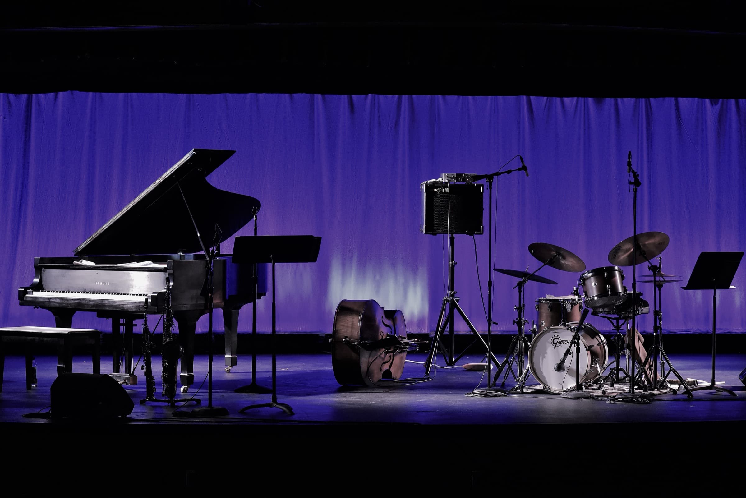 An empty stage set for a jazz group: a grand piano, an upright bass lying on its side and a drum kit, in front of a purple-lit curtain.