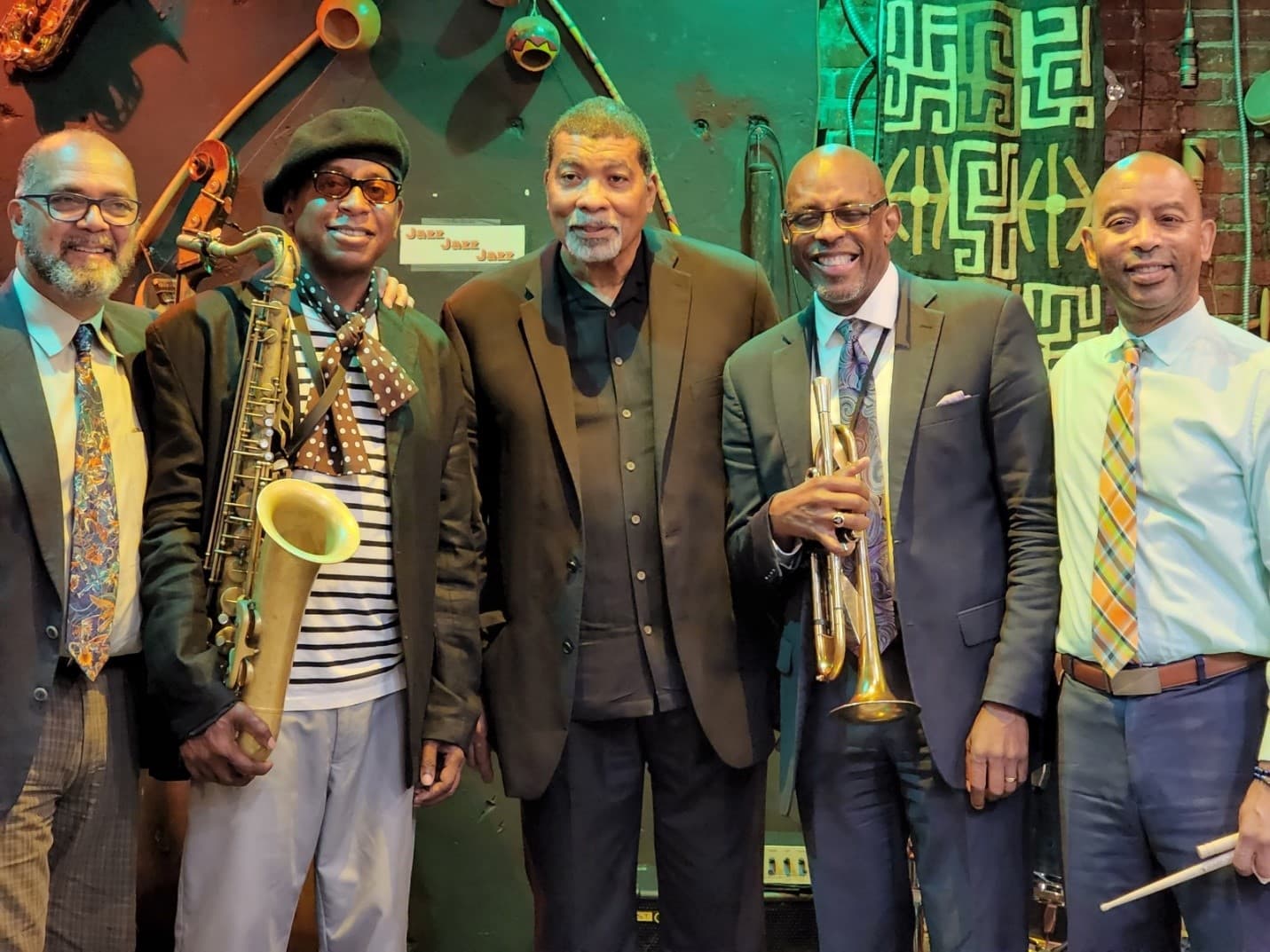 Five musicians smile for a group photo inside a jazz club. One holds a saxophone, one a trumpet, and one a pair of drumsticks.