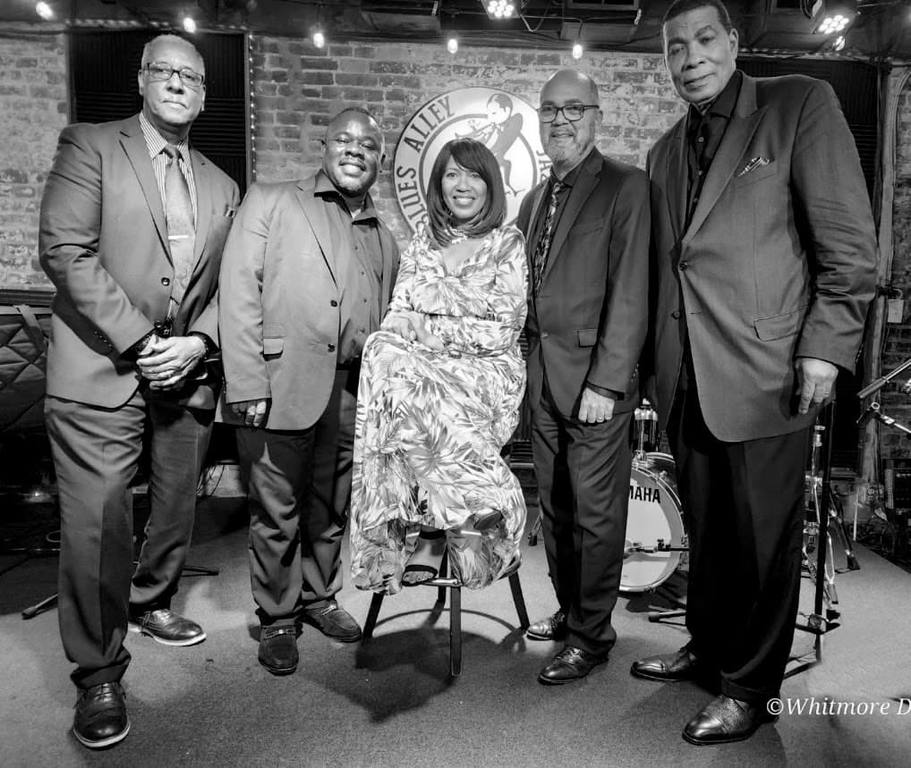 A black-and-white group portrait on the Blues Alley stage: four men in suits stand around a woman in a floral dress seated on a stool, with a drum kit behind them.