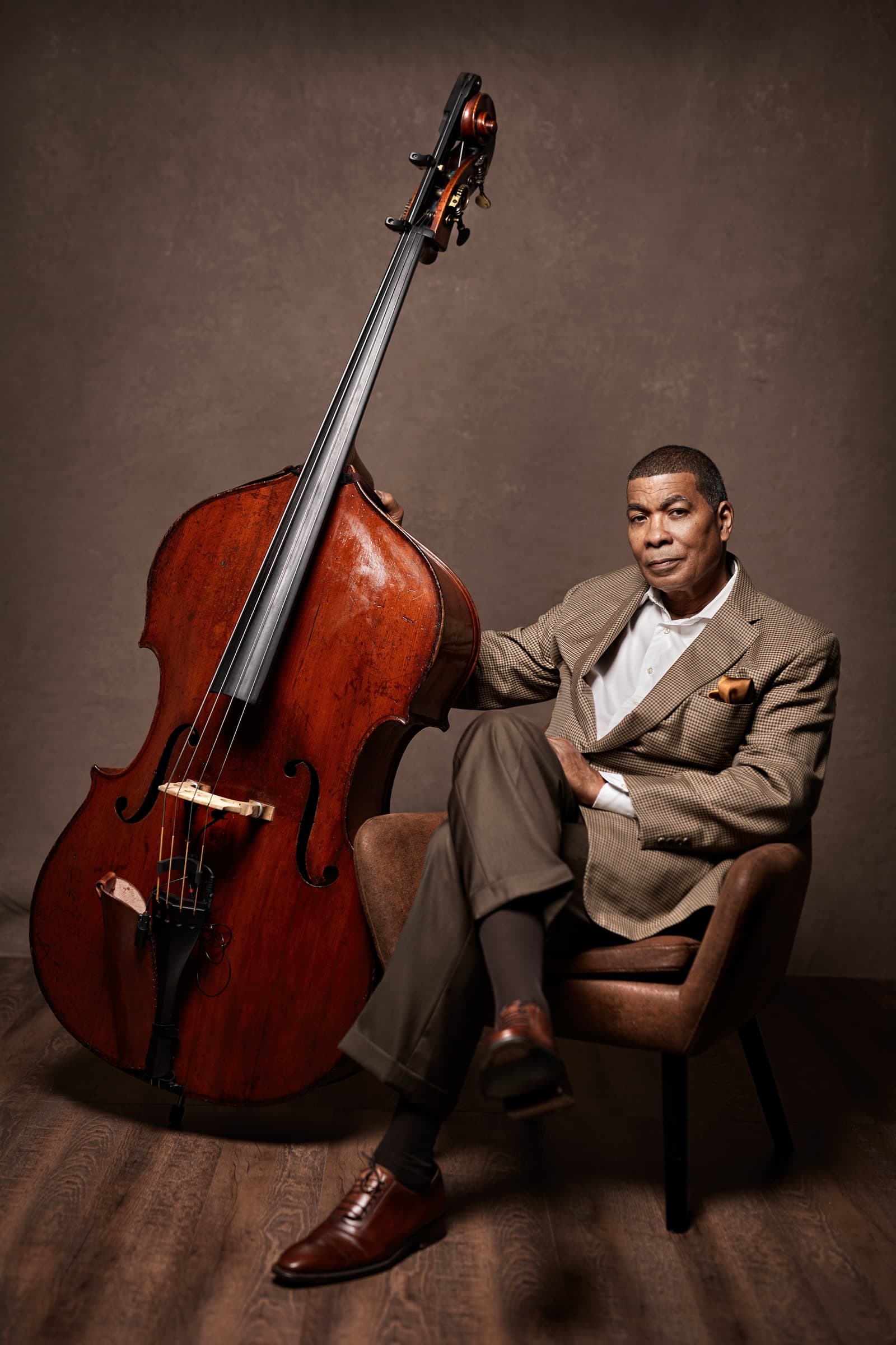 Kent Miller in a brown checked suit, seated in a chair beside his upright bass in a photo studio.