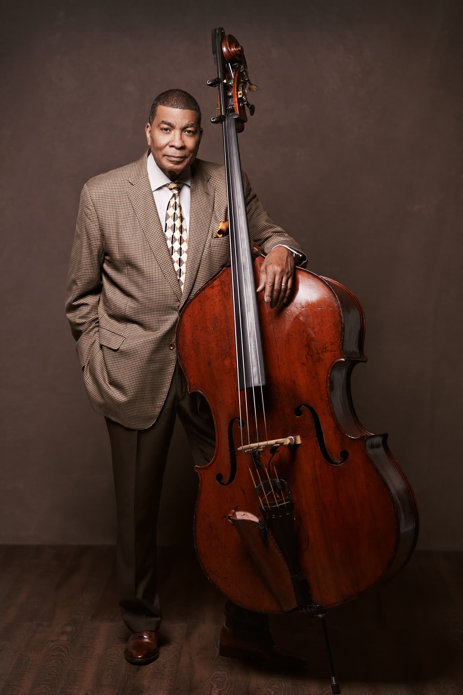 Kent Miller in a brown checked suit, standing beside his upright bass in a photo studio.