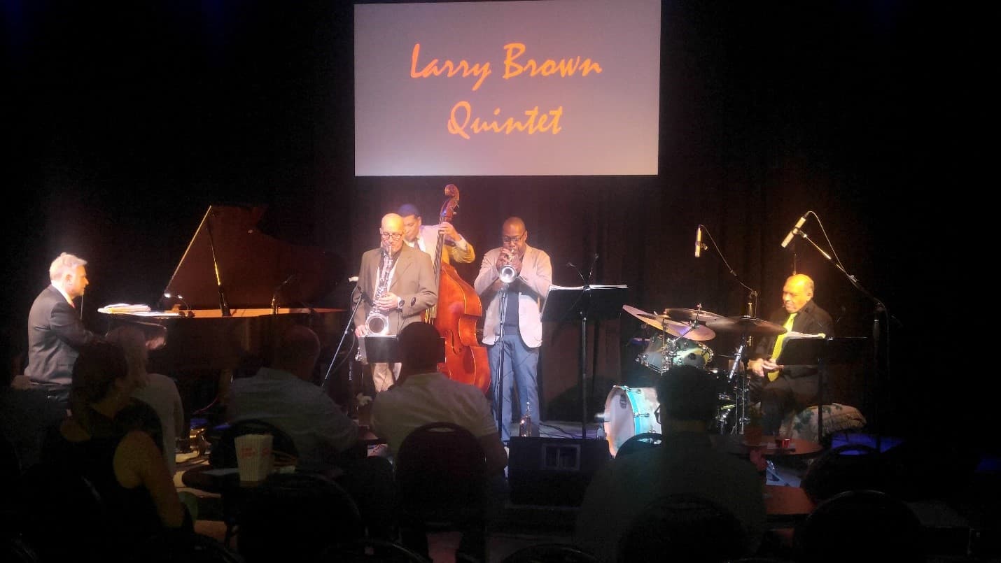 A quintet performs for a seated audience. A screen behind the band reads "Larry Brown Quintet".