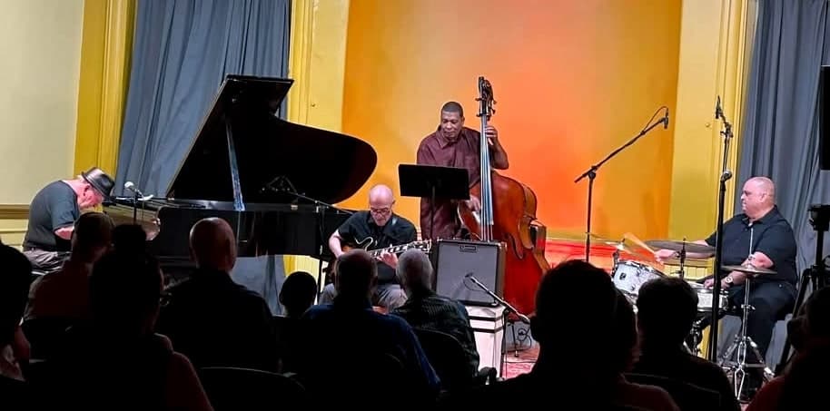 A quartet performs for a seated audience on a stage with gold walls and grey curtains: a pianist, a guitarist, a bassist and a drummer.
