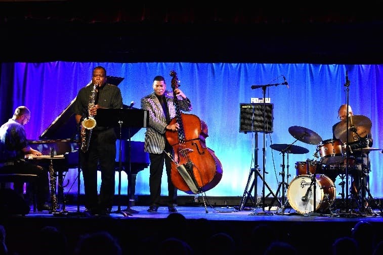 A jazz quartet performing on stage in front of a blue curtain: a pianist, a saxophonist, a bassist in a checked jacket and a drummer.