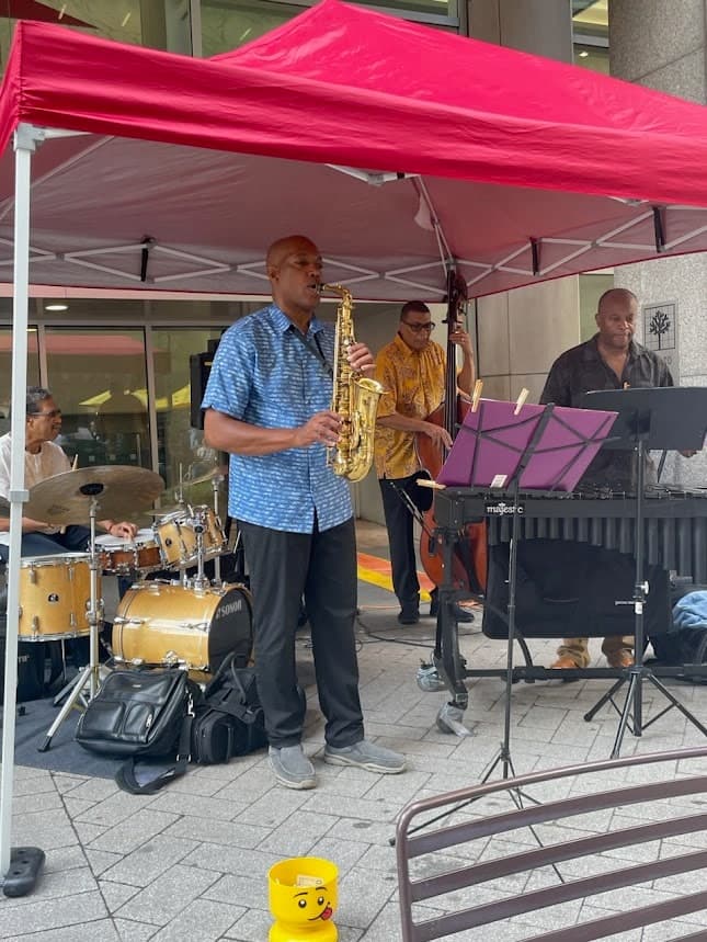 A quartet plays on a city sidewalk under a red canopy: a saxophonist in a blue shirt, a drummer, a bassist and a vibraphonist.