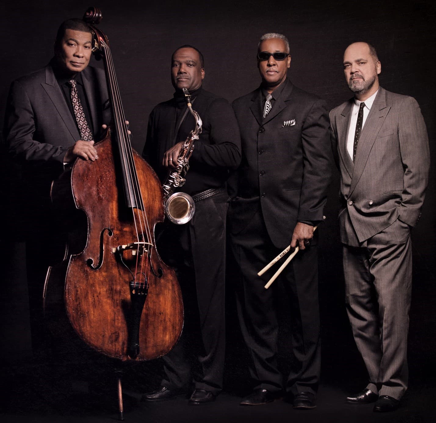 Four musicians in dark suits pose for a studio portrait. One holds an upright bass, one a saxophone, and one a pair of drumsticks.